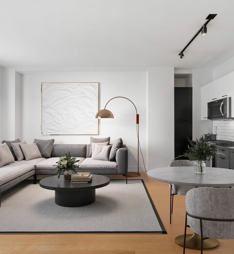 Apartments for Rent in Financial District, NY - 63 Wall Street - Living Room with Wood-Style Flooring, Area Rug, White Walls, Sectional Sofa, Dining Area, Kitchen, and Large Window.